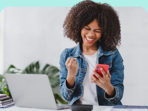 Mulher alegre comemorando assinatura do Exmed Pass enquanto segura um smartphone, com um laptop e plantas ao fundo em um ambiente iluminado.