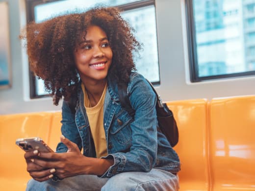 Mulher jovem e sorridente com cabelo cacheado sentada em um banco de transporte público, utilizando o app Exmed.