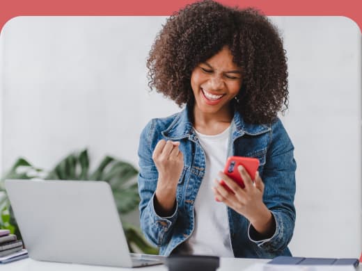 Mulher alegre comemorando assinatura do Exmed Pass enquanto segura um smartphone, com um laptop e plantas ao fundo em um ambiente iluminado.